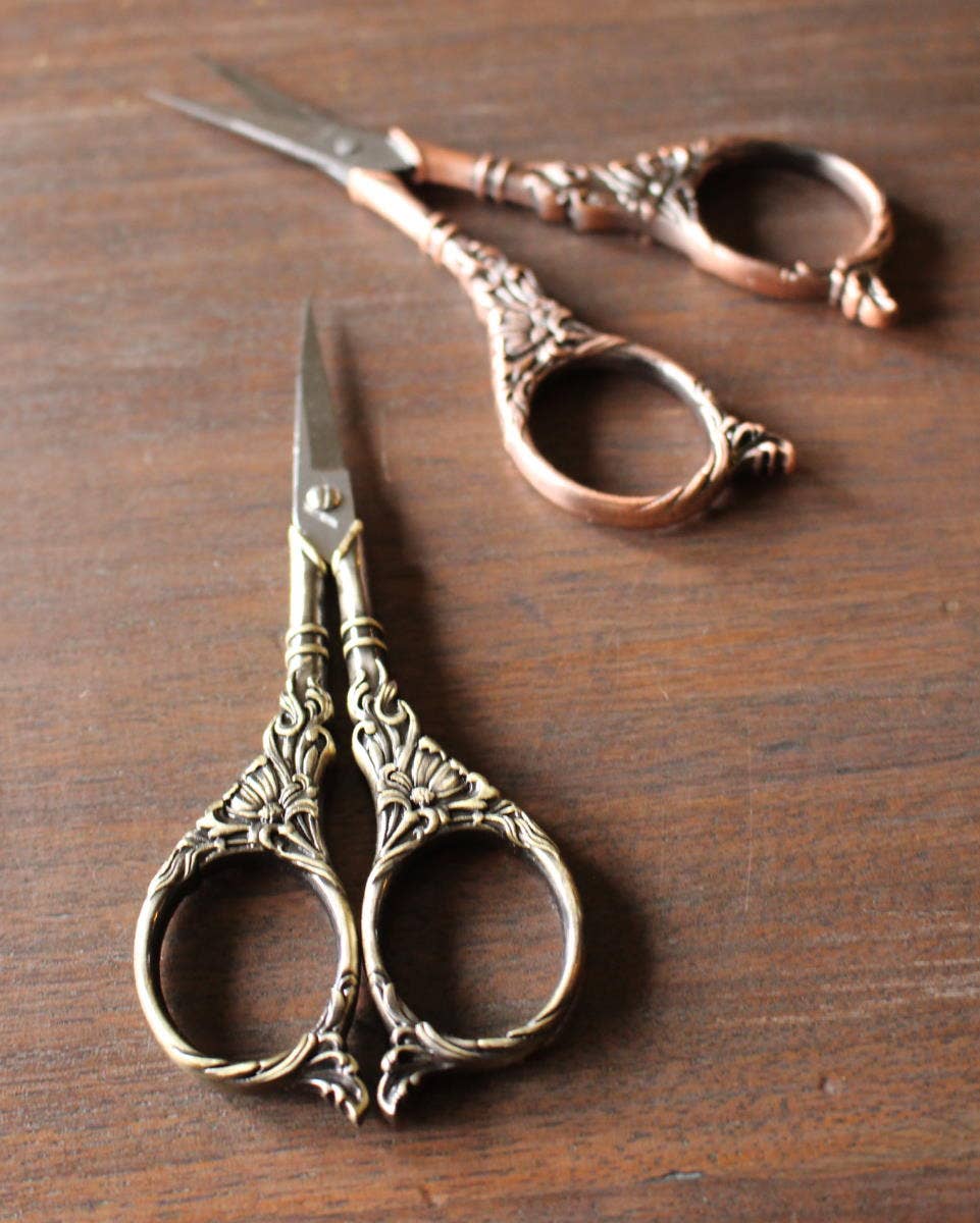 Two pairs of NNK Press Botanical Garden Scissors Antique Gold, featuring stainless steel blades and ornate botanical handles, rest on wood—one pair with an antique gold finish in front, the other with a copper finish behind.