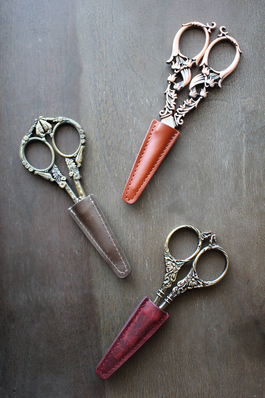 Three ornate vintage embroidery scissors with intricate handles are placed diagonally on wood, each partially sheathed in NNK Press Leather Scissor Sheaths in brown, tan, and red.