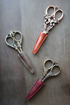 Three ornate vintage embroidery scissors with intricate handles are placed diagonally on wood, each partially sheathed in NNK Press Leather Scissor Sheaths in brown, tan, and red.
