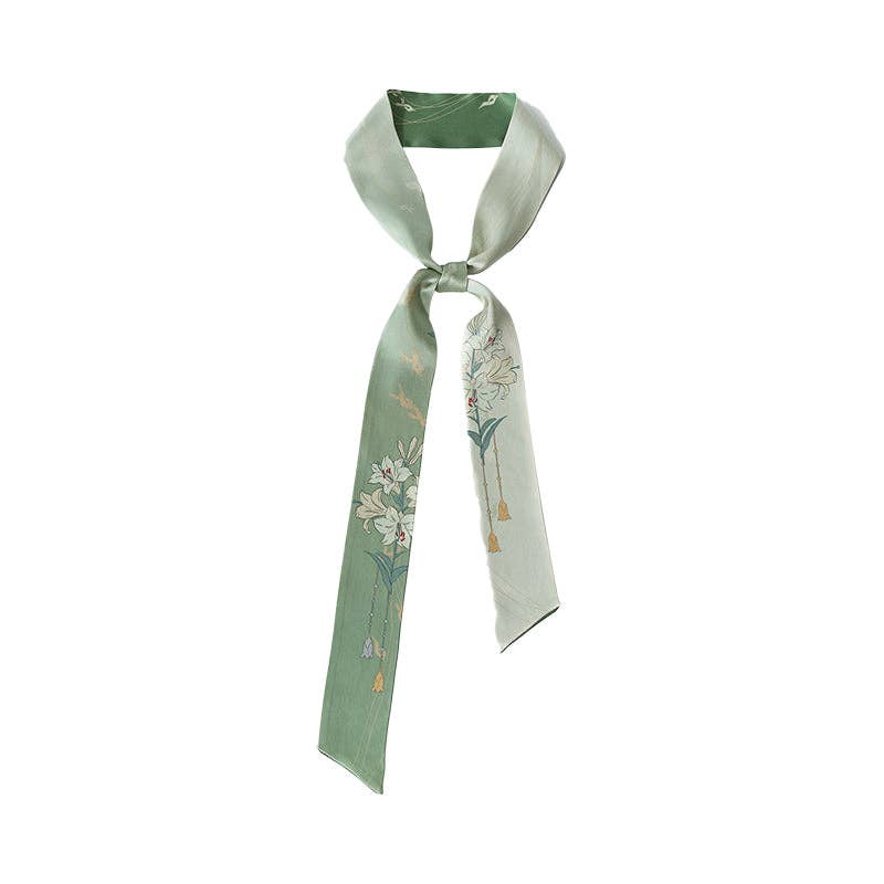 The Lily Silk Twilly Scarf: Green by Lost Pattern NYC features white floral designs and gold tassel accents, offering a chic touch as a hair scarf or an elegant addition to any gift box.