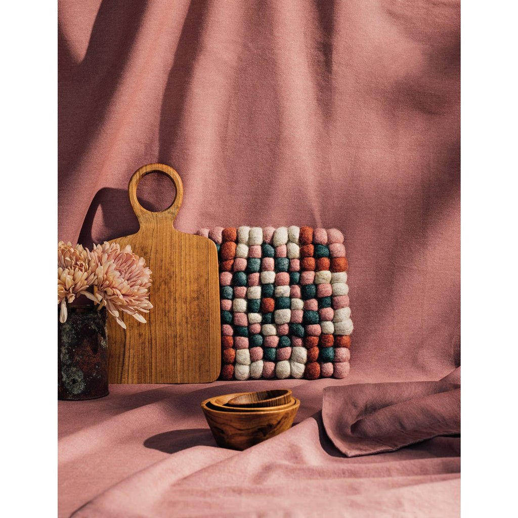 A wooden cutting board, a small wooden bowl, the Danica Heirloom Terra Dot Trivet, and a vase with pink flowers are arranged on a pink cloth background.