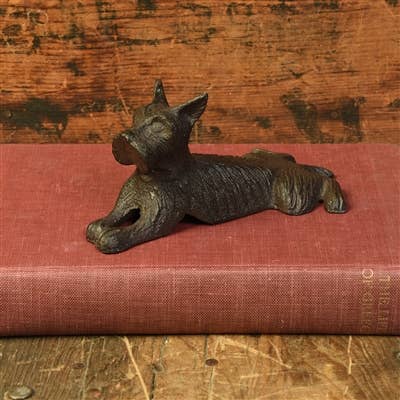 The HomArt Terrier Door Wedge, Cast Iron in Antique Brown, shaped like a dog, rests atop a closed red book on a rustic wooden background.