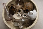 A round bowl holds assorted bottles, including the Indaba Trading Recycled Glass Bottle Vase in clear glass, alongside vintage-style ceramic and glass vases in earthy tones, seen from above on a light fabric surface.