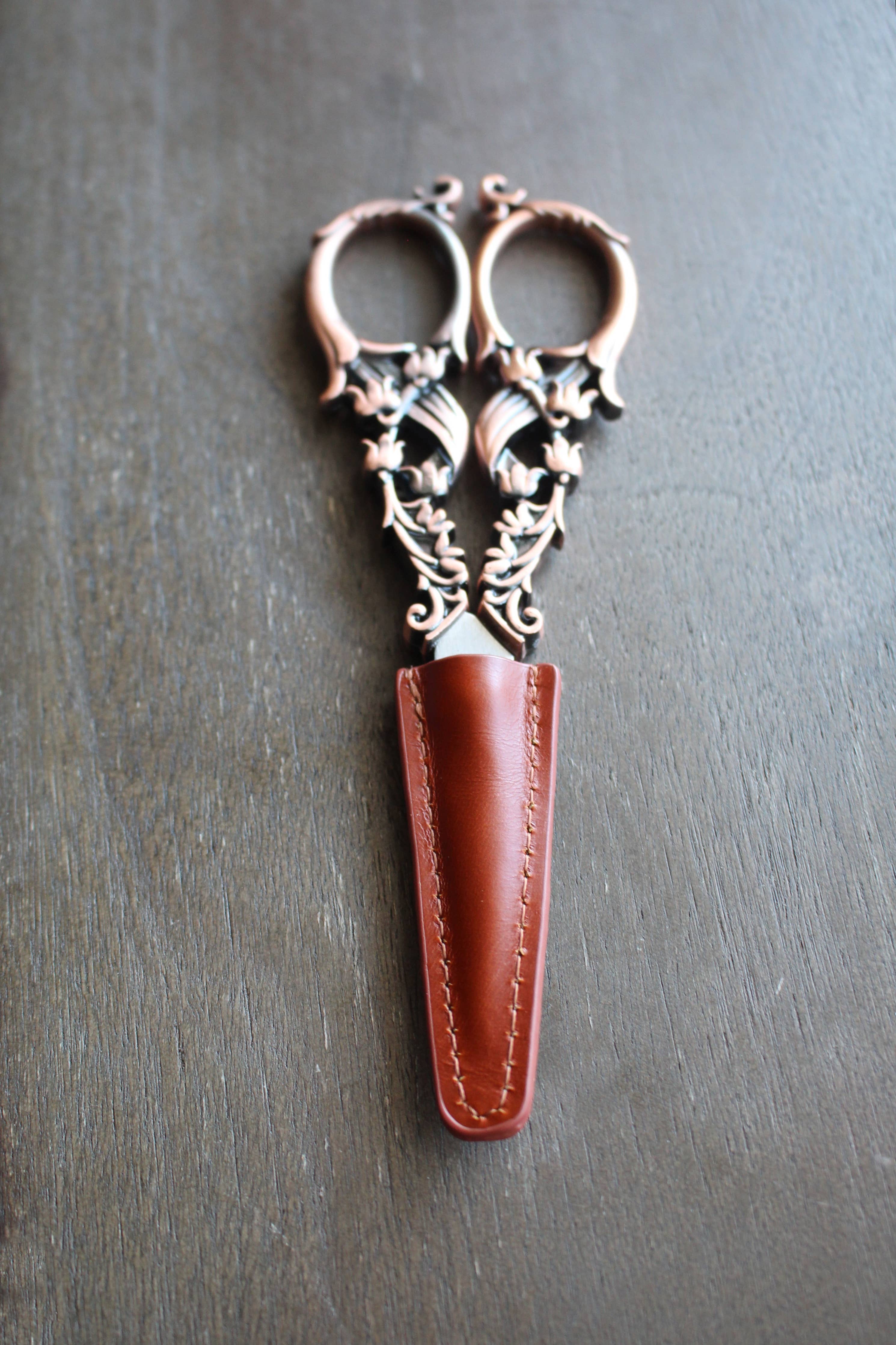 A pair of ornate embroidery scissors rest on a wooden surface, partially sheathed in the NNK Press Leather Scissor Sheath.