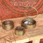 The HomArt Josephine Mirrored Box in small round brass rests on a carved wooden surface, open to reveal treasures inside. Beside it are two other ornate boxes, with a red patterned textile hanging in the background.