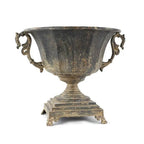 The Distressed Black Iron Urn by Vagabond Vintage features ornate handles and paw feet on a square base, with a weathered finish and shown against a white background.