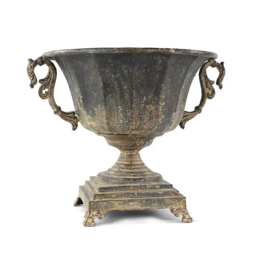 The Distressed Black Iron Urn by Vagabond Vintage features ornate handles and paw feet on a square base, with a weathered finish and shown against a white background.