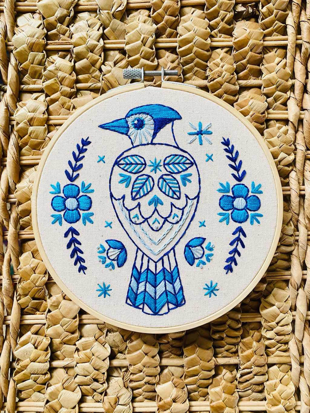The Blue Jay Complete Embroidery Kit by Hook, Line & Tinker features a blue and white embroidered bird among floral and leaf patterns on straw fabric—perfect for beginners who love intricate blue designs.