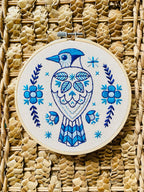 The Blue Jay Complete Embroidery Kit by Hook, Line & Tinker features a blue and white embroidered bird among floral and leaf patterns on straw fabric—perfect for beginners who love intricate blue designs.