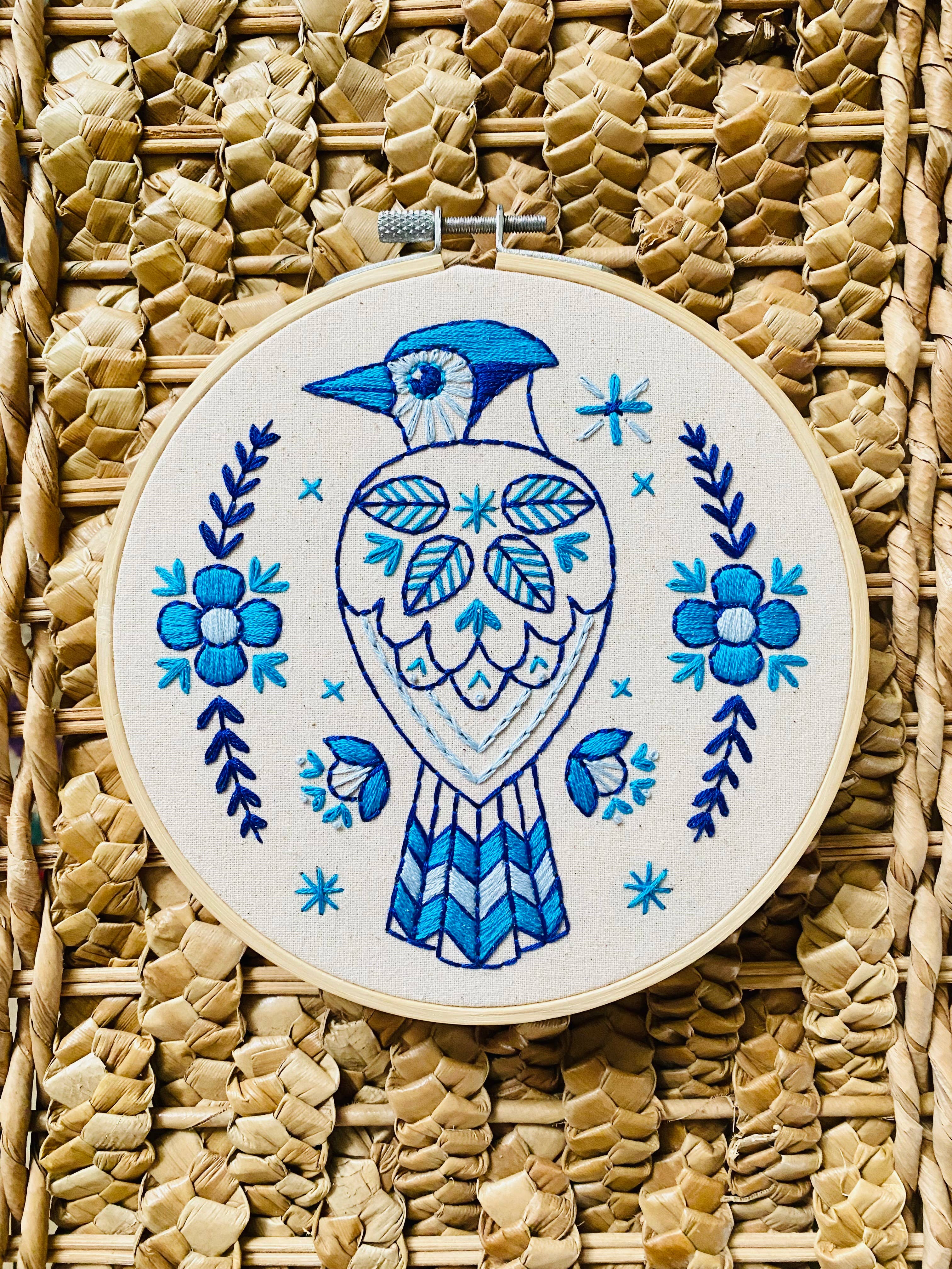 The Blue Jay Complete Embroidery Kit by Hook, Line & Tinker features a blue and white embroidered bird among floral and leaf patterns on straw fabric—perfect for beginners who love intricate blue designs.