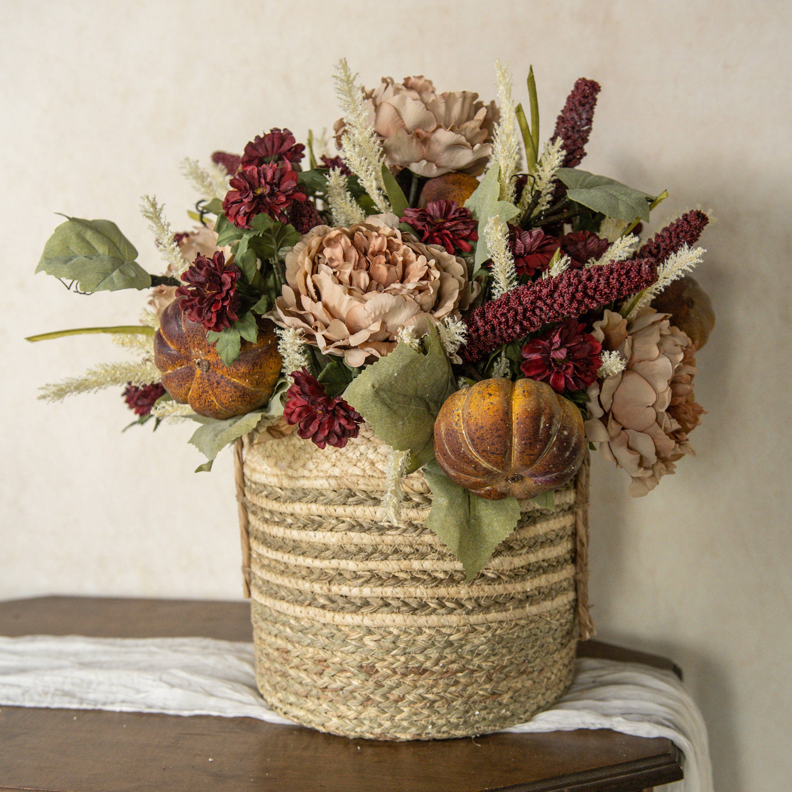A 31" BURGUNDY SORGHUM SPRAY by Ragon House is arranged with autumn florals, pumpkins, peonies, and green leaves in a rustic basket on a wooden surface against a neutral background.