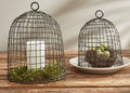 The Park Designs Wire Bell Cloches - Set of 2 feature elegant circular handles and display a candle with greenery under one cloche, while the other shelters a nest with eggs on stacked white plates, all arranged on a wooden surface.