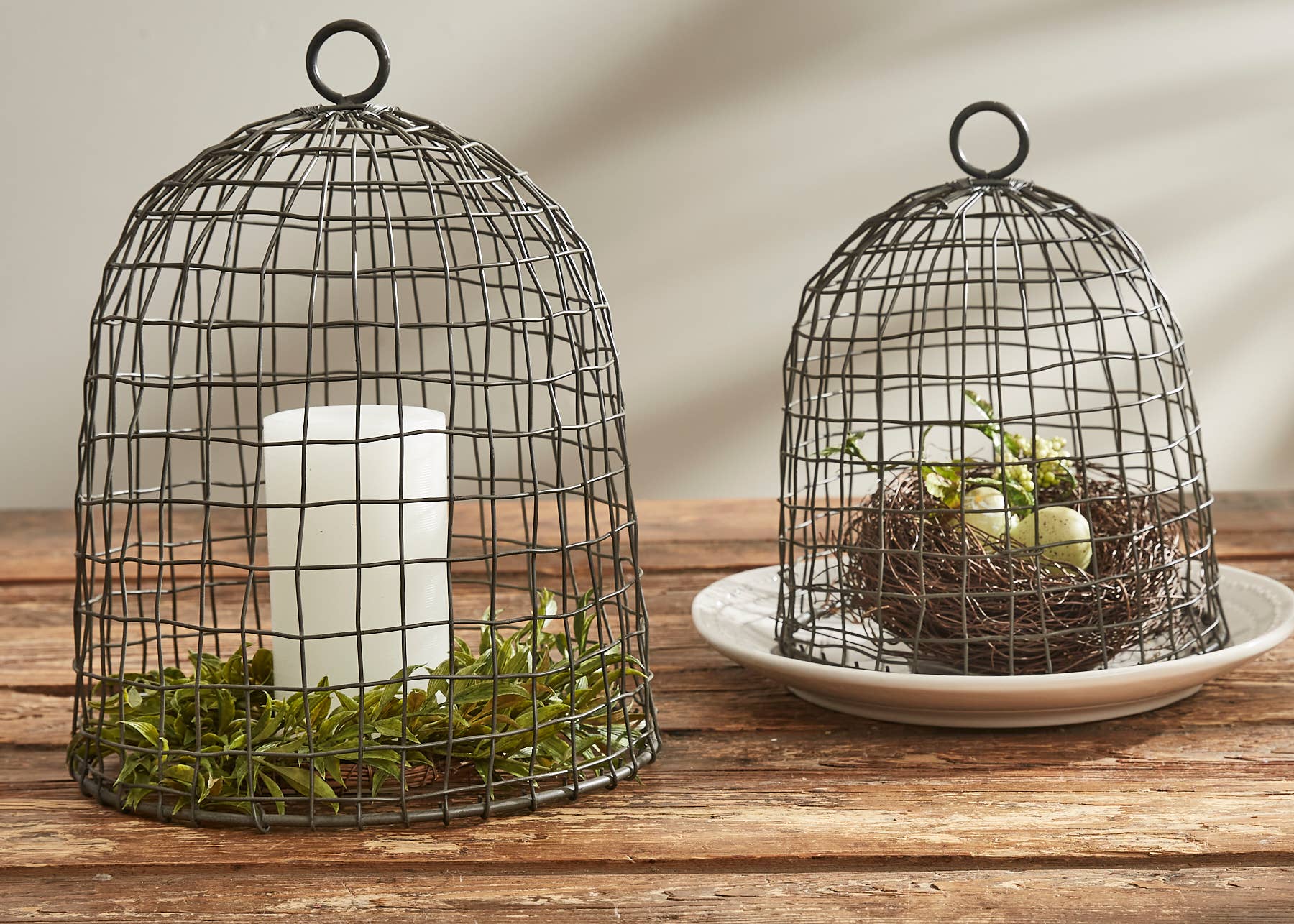 The Park Designs Wire Bell Cloches - Set of 2 feature elegant circular handles and display a candle with greenery under one cloche, while the other shelters a nest with eggs on stacked white plates, all arranged on a wooden surface.