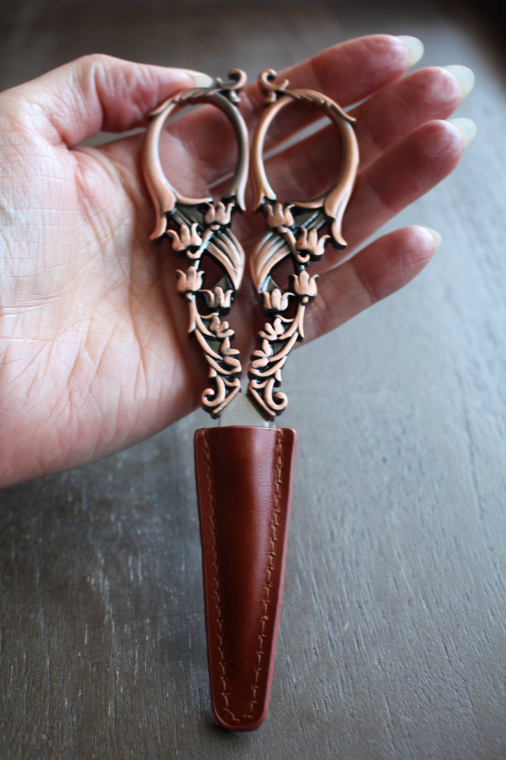 A hand holds a pair of ornate bronze embroidery scissors with leaf designs, partly sheathed in the NNK Press Leather Scissor Sheath, shown against a wooden background.