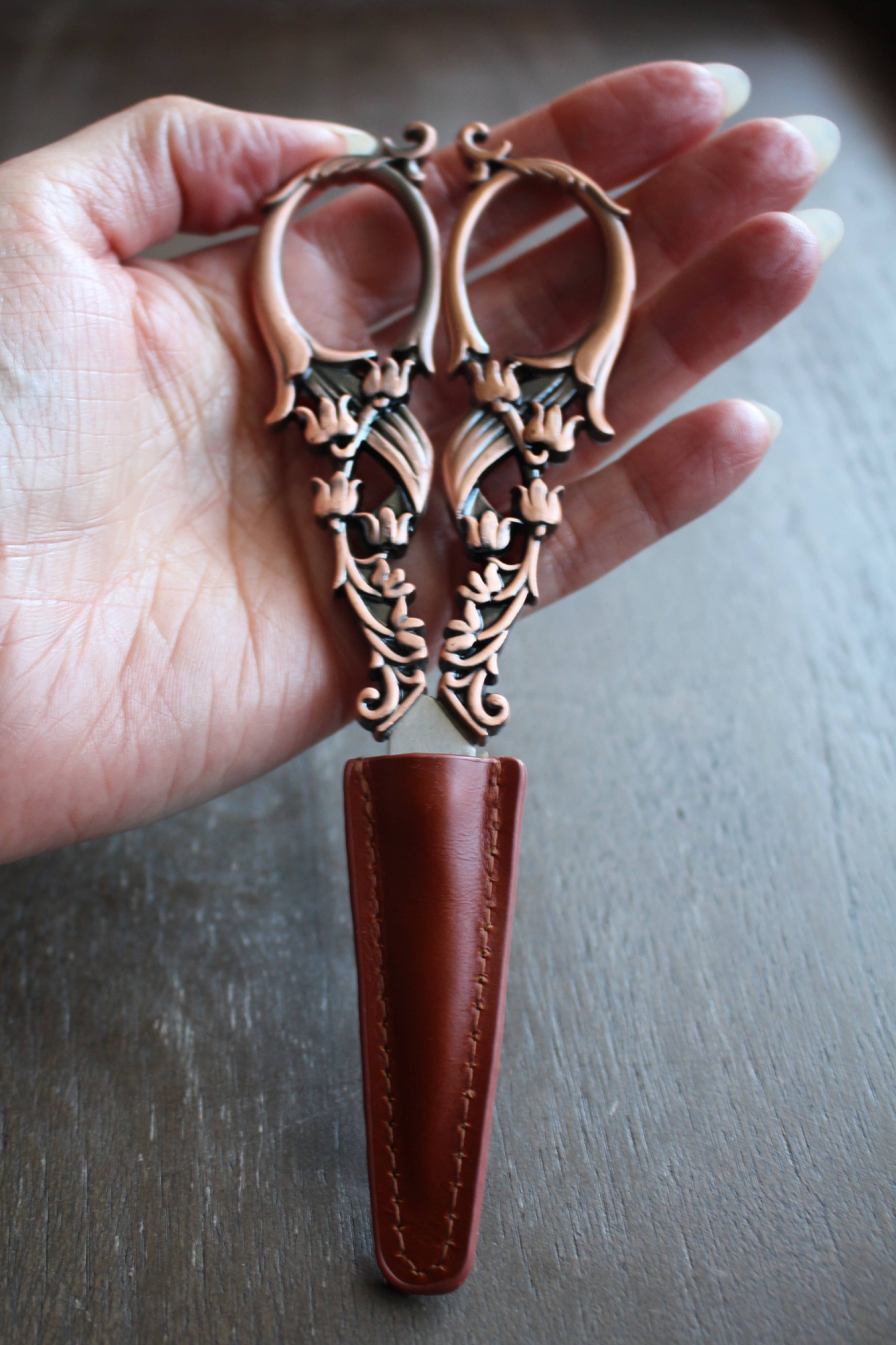 A hand holds a pair of ornate bronze embroidery scissors with leaf designs, partly sheathed in the NNK Press Leather Scissor Sheath, shown against a wooden background.