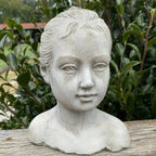 The Cement Girl Statue by Vagabond Vintage features a delicately crafted young girl with tied-back hair, perfect for adding an elegant touch to your garden decor amidst greenery.