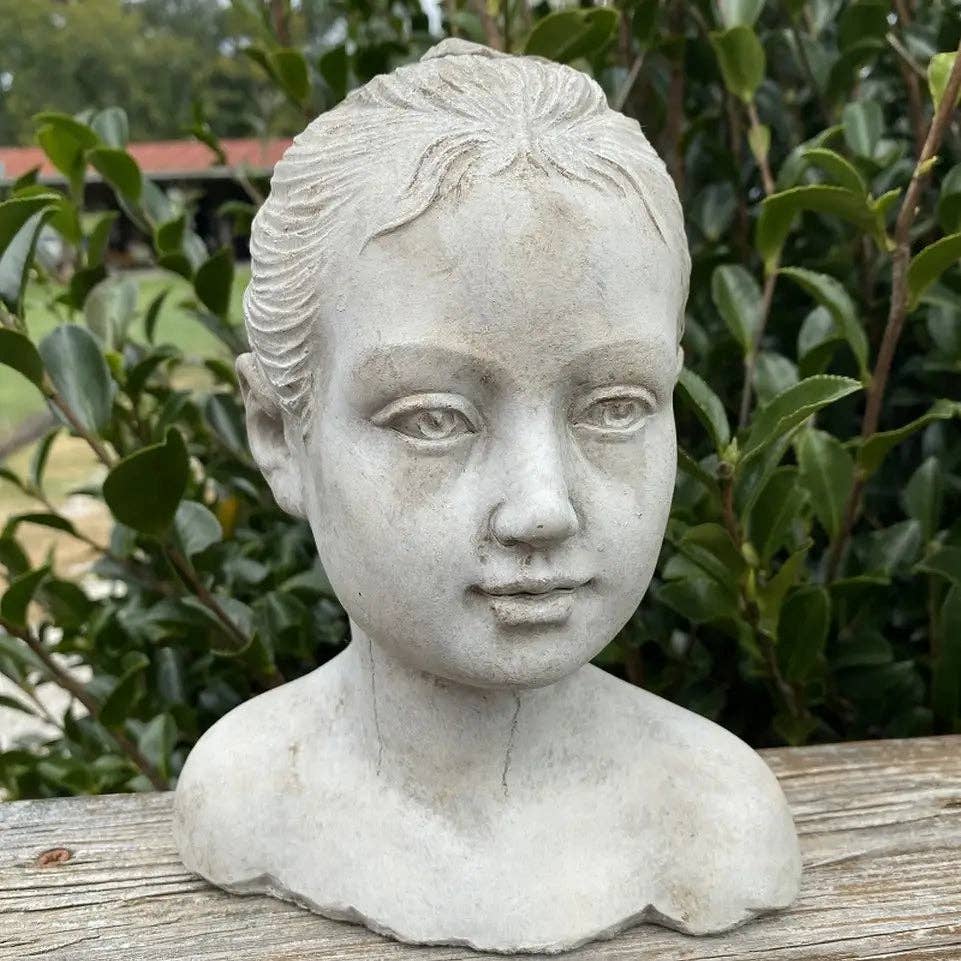 The Cement Girl Statue by Vagabond Vintage features a delicately crafted young girl with tied-back hair, perfect for adding an elegant touch to your garden decor amidst greenery.