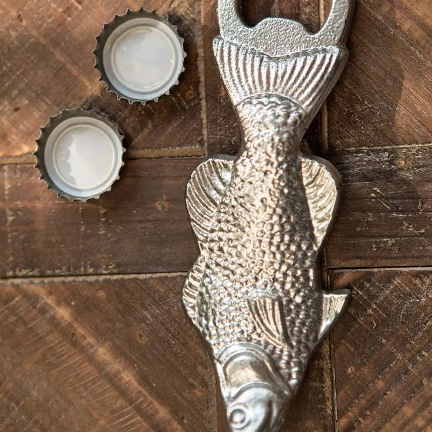 The Vagabond Vintage Metal Fish Bottle Opener, designed for comfort and ease of use, rests on a wooden surface beside two opened bottle caps.