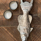 The Vagabond Vintage Metal Fish Bottle Opener, designed for comfort and ease of use, rests on a wooden surface beside two opened bottle caps.
