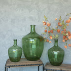A 16" GREEN GLASS BOTTLE from Ragon House is displayed with two smaller green vases on metal and wood stands against a textured gray wall, with orange lantern flowers creating a striking centerpiece in the rightmost vase.