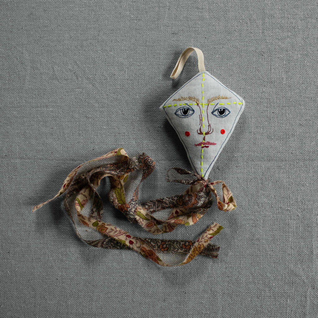 The Skippy Cotton Flying Kite - Cotton & Lavender Filled Ornament, Scented, features a small, diamond shape with a painted face and red cheeks, and a long patterned fabric tail, resting on a textured gray surface.