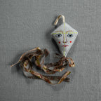 The Skippy Cotton Flying Kite - Cotton & Lavender Filled Ornament, Scented, features a small, diamond shape with a painted face and red cheeks, and a long patterned fabric tail, resting on a textured gray surface.