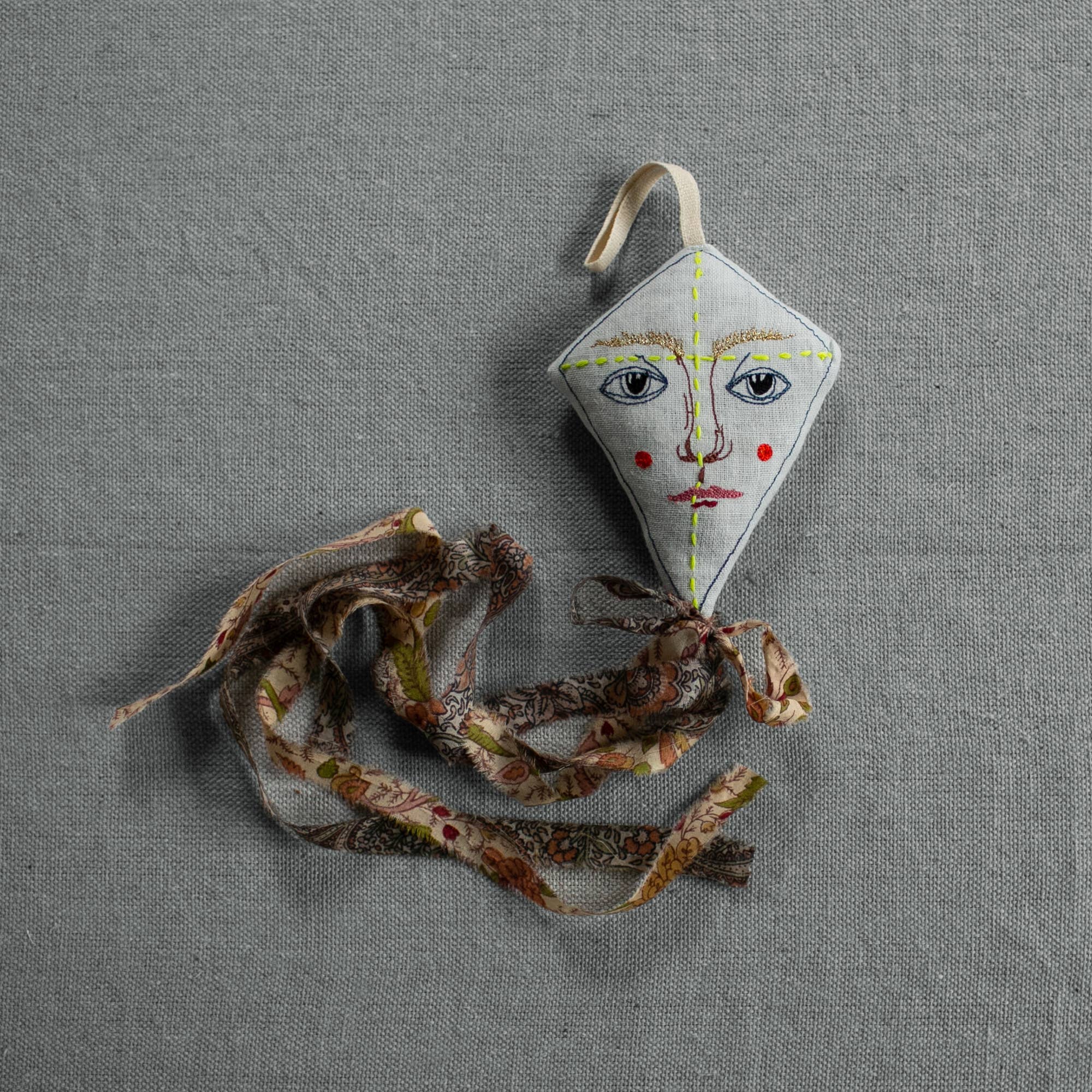 The Skippy Cotton Flying Kite - Cotton & Lavender Filled Ornament, Scented, features a small, diamond shape with a painted face and red cheeks, and a long patterned fabric tail, resting on a textured gray surface.