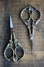 The NNK Press Feathered Friends Scissors in Antique Gold feature ornate bird and floral designs, pointed stainless steel blades, and an antique finish, shown resting on a rustic wooden surface.