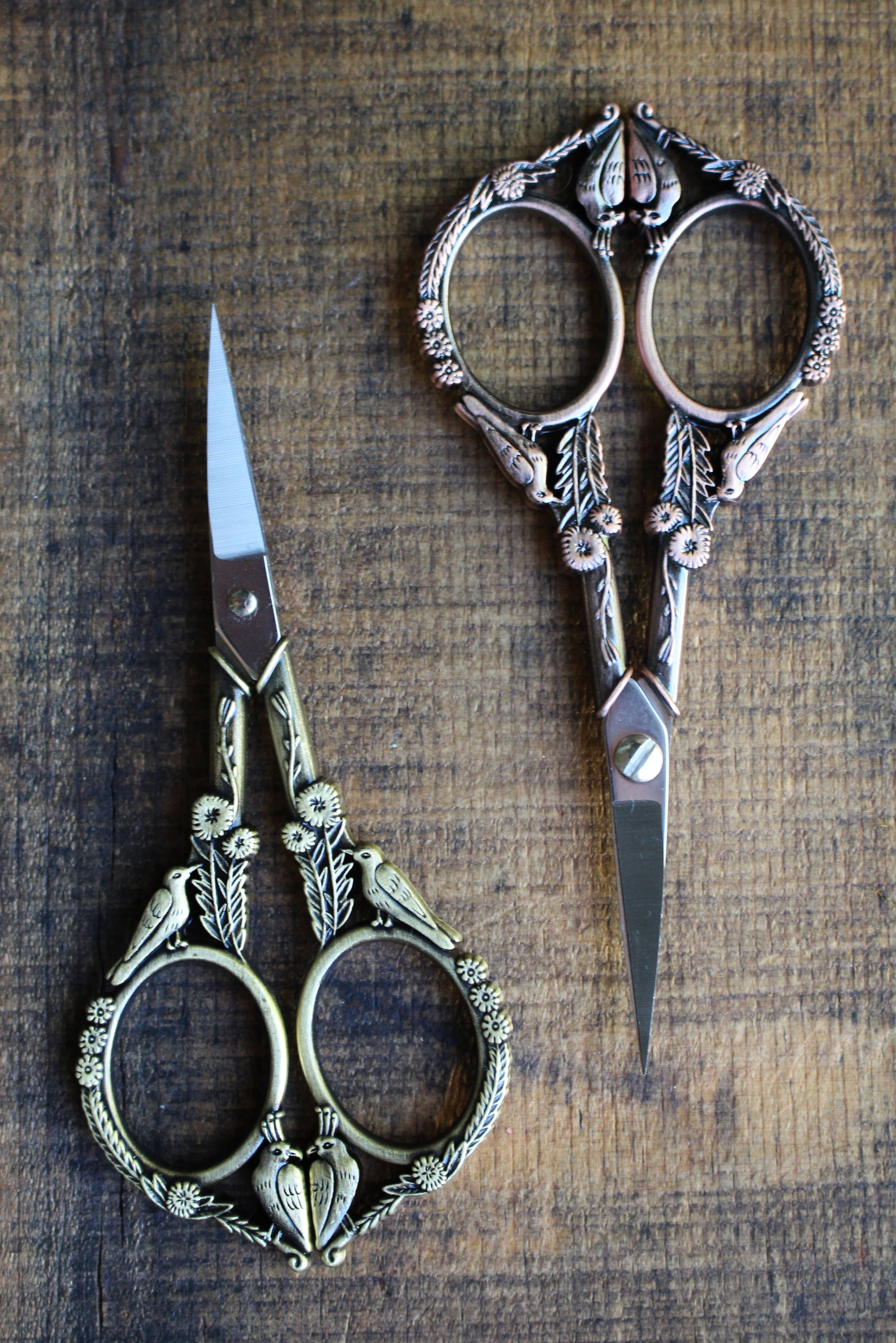 The NNK Press Feathered Friends Scissors in Antique Gold feature ornate bird and floral designs, pointed stainless steel blades, and an antique finish, shown resting on a rustic wooden surface.