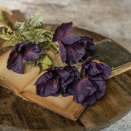 A 22" Artificial Dark Purple Poppy Bush by Ragon House lies on the yellowed pages of an old book atop rustic wood, with a closed book nearby, creating a vintage and nostalgic atmosphere.