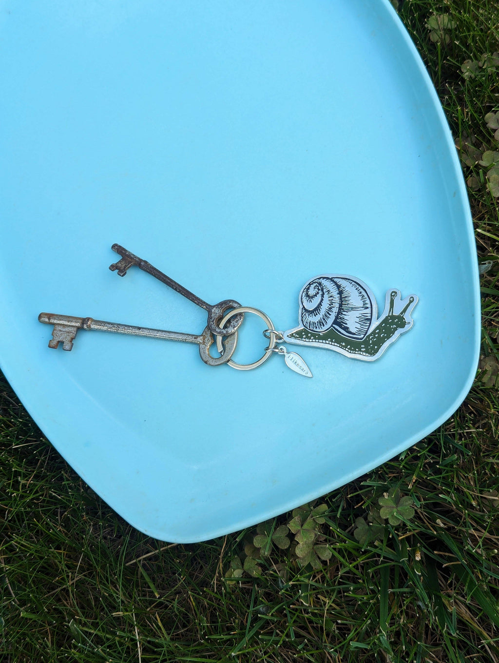 A pair of vintage-style keys and the ilikesara art + goods metal enamel snail keychain rest on a light blue plate placed on green grass, with clover leaves peeking around the edges.