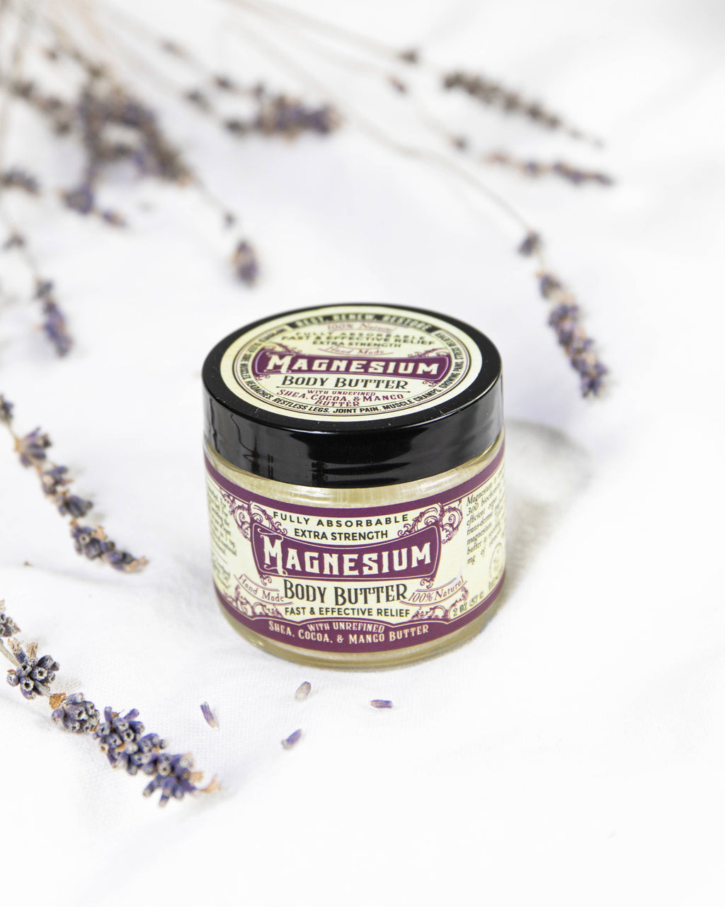 A jar of Roots And Leaves Magnesium Body Butter with a vintage-style label rests on a white surface, surrounded by dried lavender, emphasizing its role as a sleep aid and for boosting magnesium absorption.