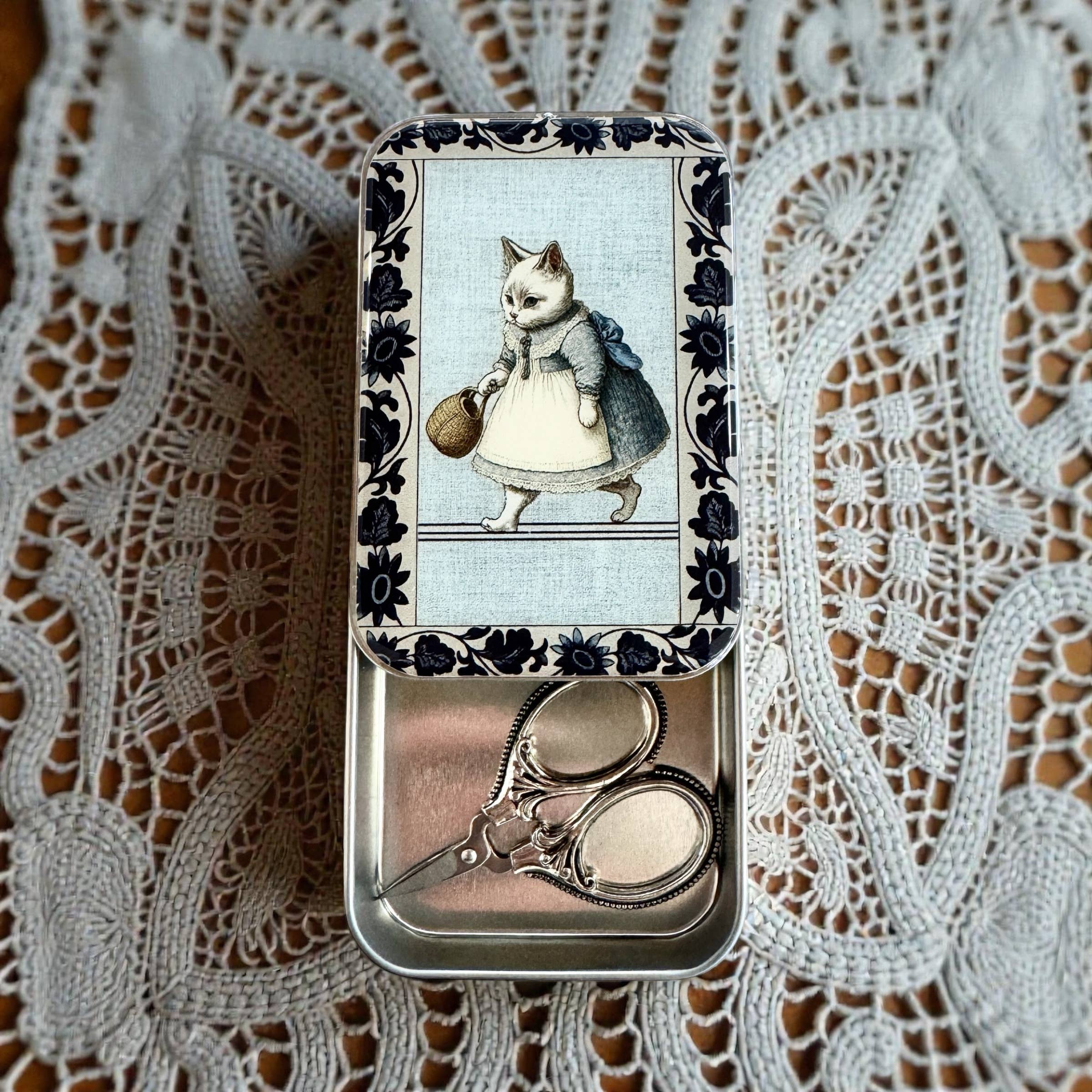 The Firefly Notes Kitty notions tin: Small is a metal tin with embroidery scissors inside, resting on lace. The lid features Business Cat in a blue dress with a basket, surrounded by a decorative blue border.