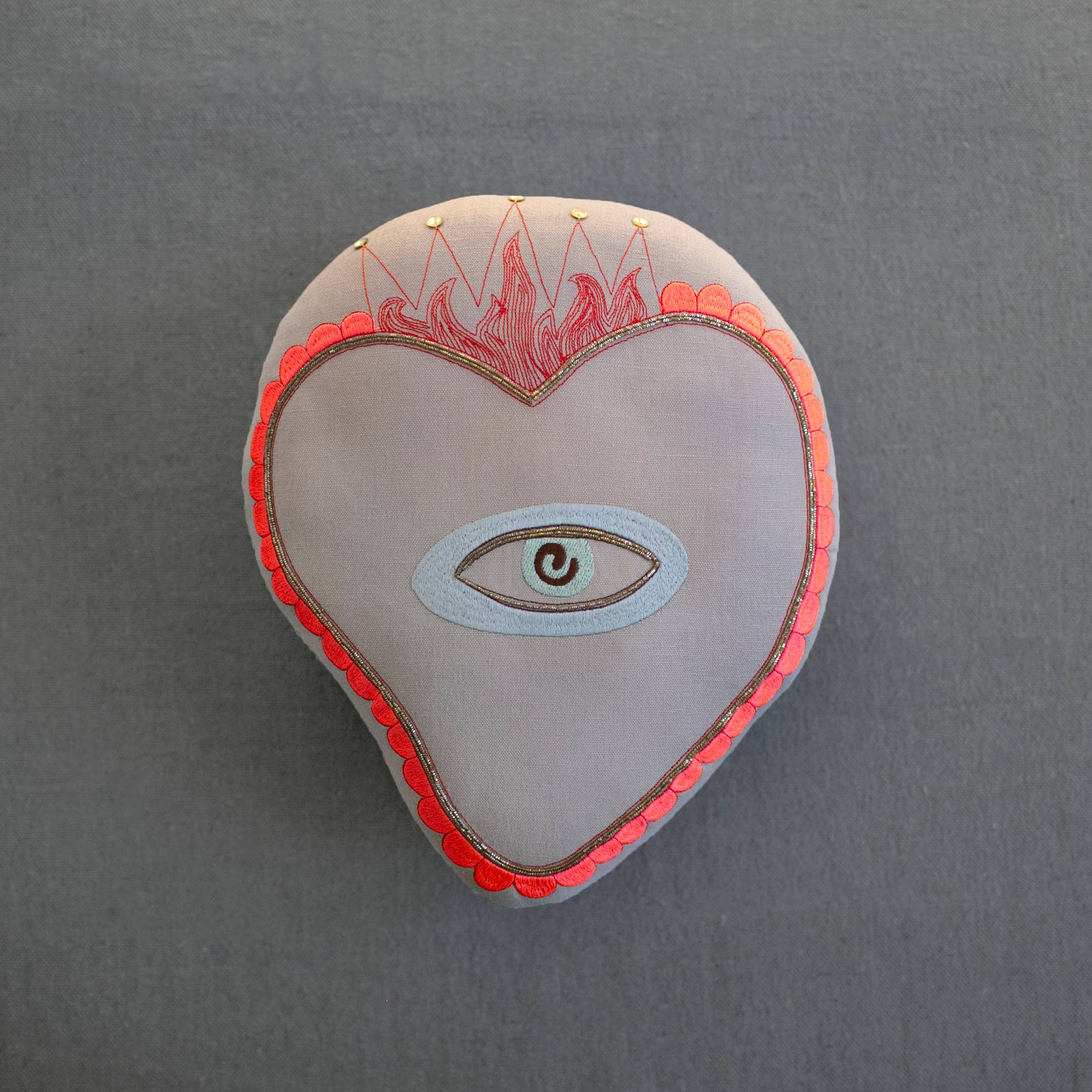 The Sacred Heart Pillow by Skippy Cotton is a handmade, cotton-filled cushion with red trim, an embroidered central eye, and a flame motif—ideal for whimsical decor. Shown on a plain gray background.