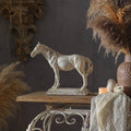 The Rustic Horse Sculpture by Vagabond Vintage sits on a wooden table with a beige cloth, candle, bird figurine, and dried pampas grass in a rustic vase for a cozy farmhouse decor look.