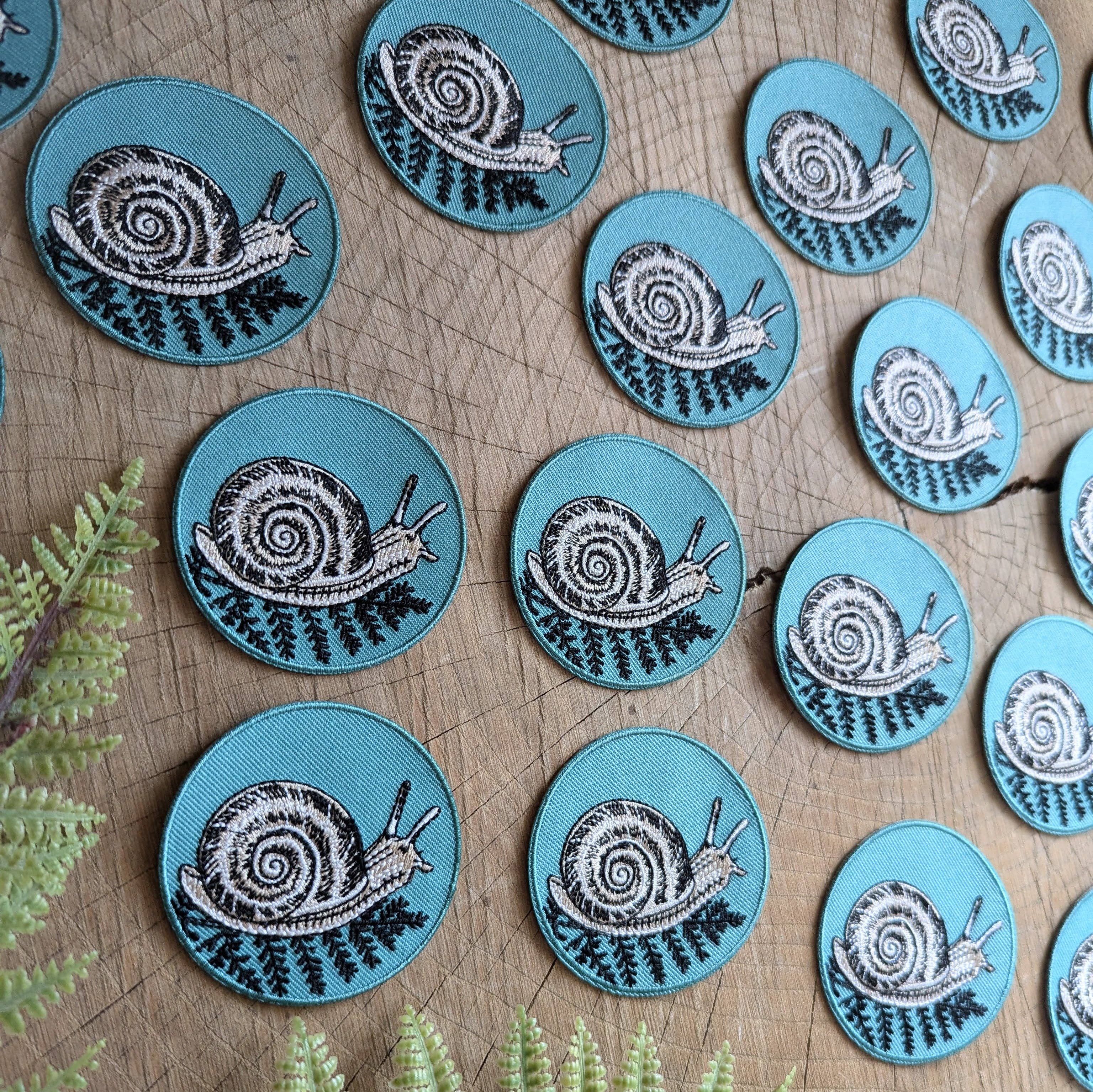 Several ilikesara | art + goods 2.5" round teal snail with fern iron-on patches, featuring black and white snail illustrations, are displayed on wood with green fern leaves, sold without backer card or packaging.