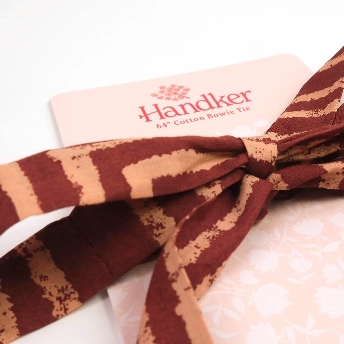 A pink and brown striped Sammie Pink Bowie Tie by Handker Bandanas knotted and resting on a branded product card.