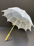 The Parasol - White by Peking Handicraft is a Battenburg Victorian parasol with intricate lace, light wooden handle and frame, displayed open against a dark background—an elegant accessory for any occasion.