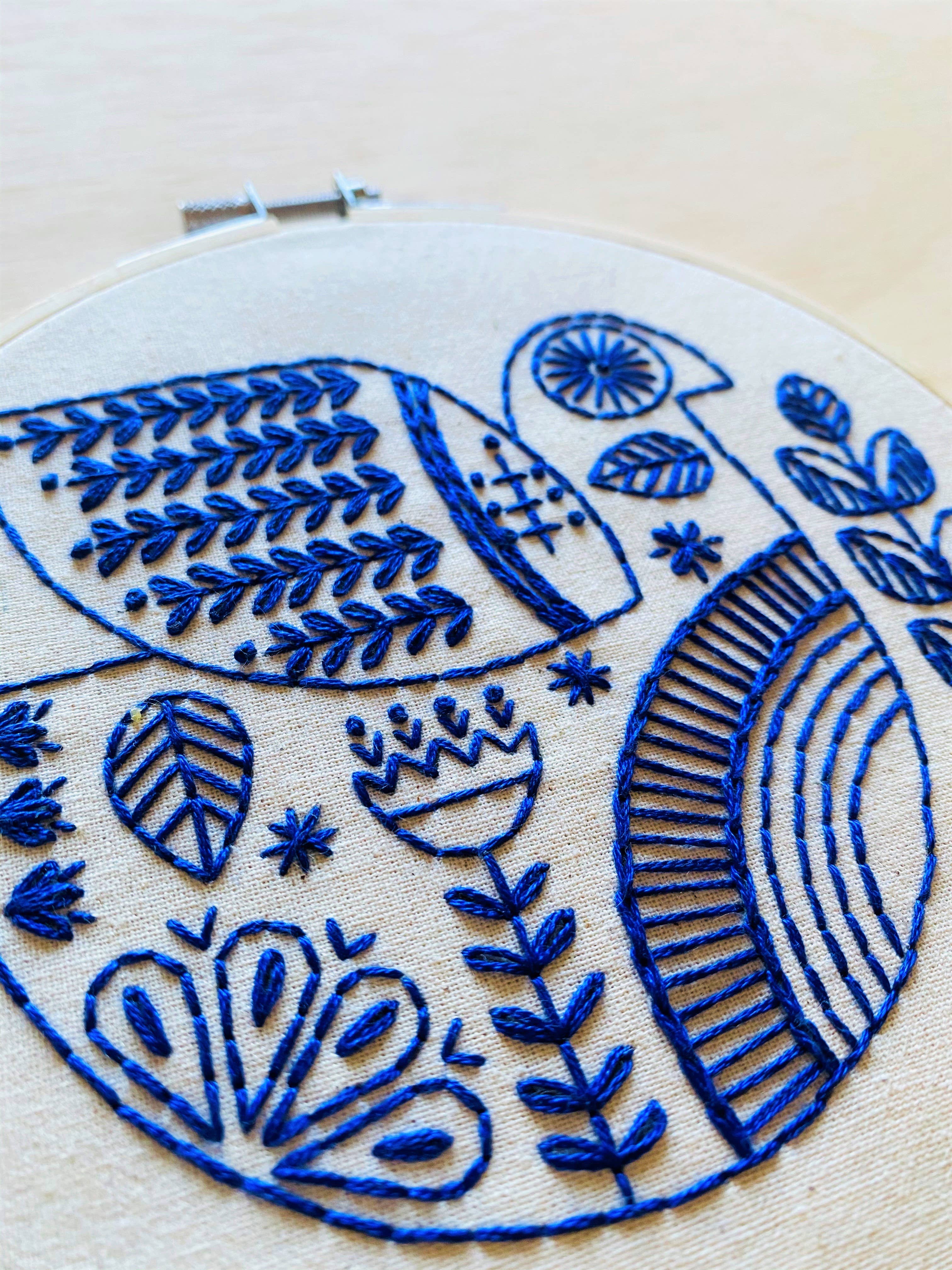 A close-up of the Hygge Dove Complete Embroidery Kit by Hook, Line & Tinker Embroidery Kits shows blue thread creating botanical and geometric patterns, such as leaves, flowers, and arcs, stitched on white fabric in an embroidery hoop.