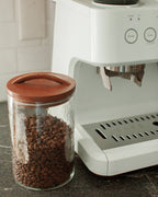 A Handblown Hammered Glass Canisters: Blush / Medium by Creative Women, filled with coffee beans and topped with a hand-carved wooden lid, sits next to a white espresso machine on a dark countertop.