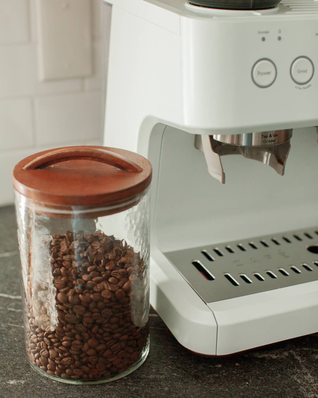 A Handblown Hammered Glass Canisters: Blush / Medium by Creative Women, filled with coffee beans and topped with a hand-carved wooden lid, sits next to a white espresso machine on a dark countertop.