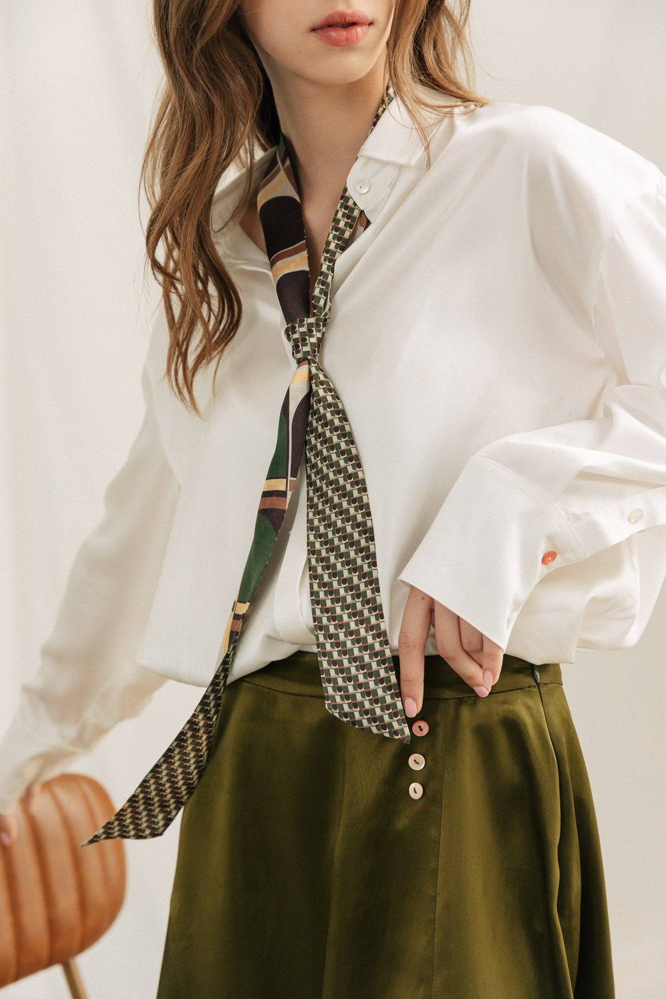 A woman stands with one hand on her hip, wearing a loose white button-up, an olive green skirt with buttons, and the Lost Pattern NYC Silk Skinny Scarf - Green styled in her hair. Her wavy hair partially covers her face.