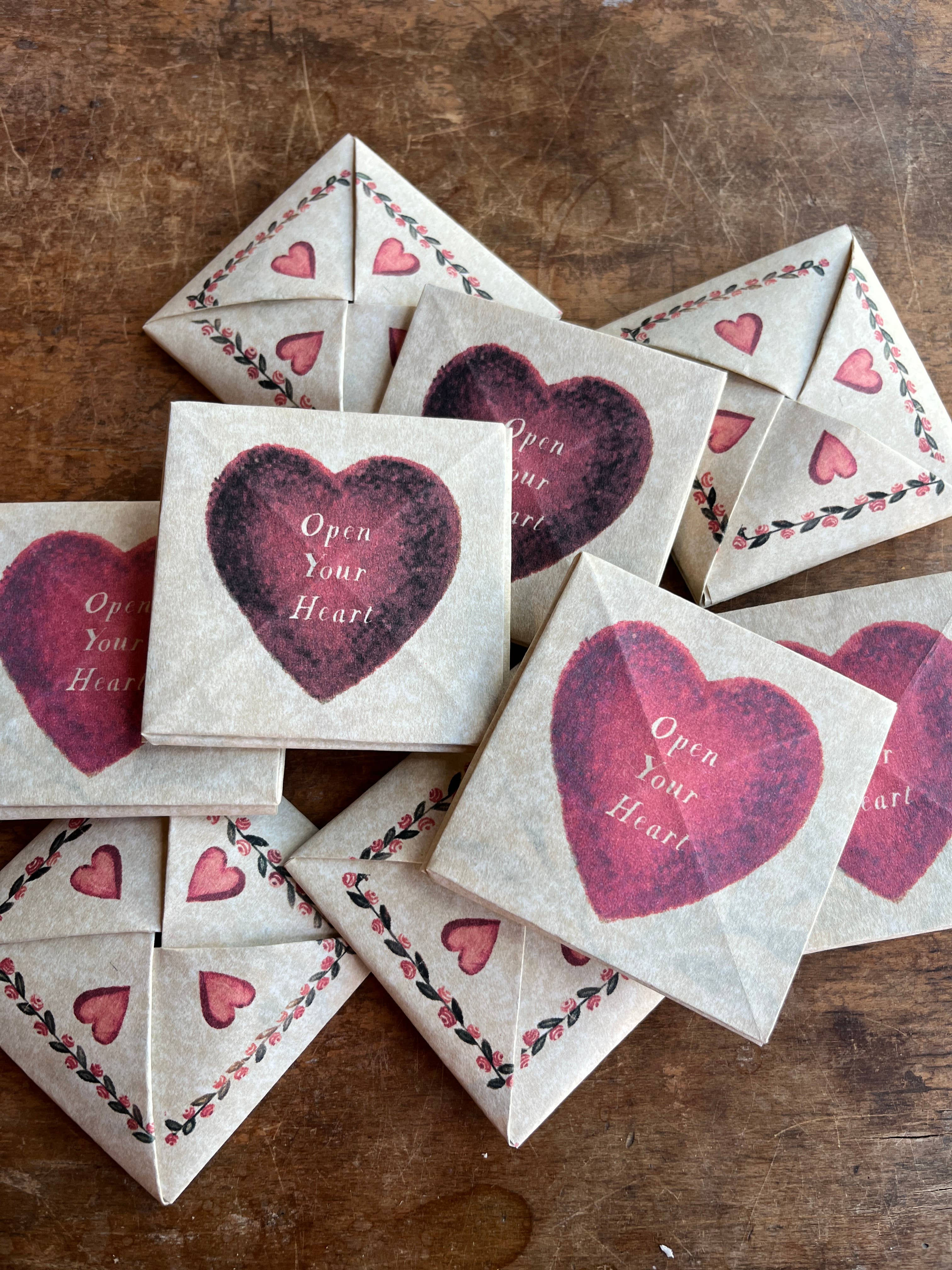 The Victorian Valentine Puzzle: Romantic by Parcel features square paper envelopes on creamy white cardstock, adorned with large red hearts, "Open Your Heart" text, and heart-vine borders, displayed on a wooden surface.