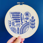 A hand holds the Hygge Dove Complete Embroidery Kit by Hook, Line & Tinker, featuring a blue and white stylized bird, leaves, and geometric patterns on an embroidery hoop against a solid blue background.