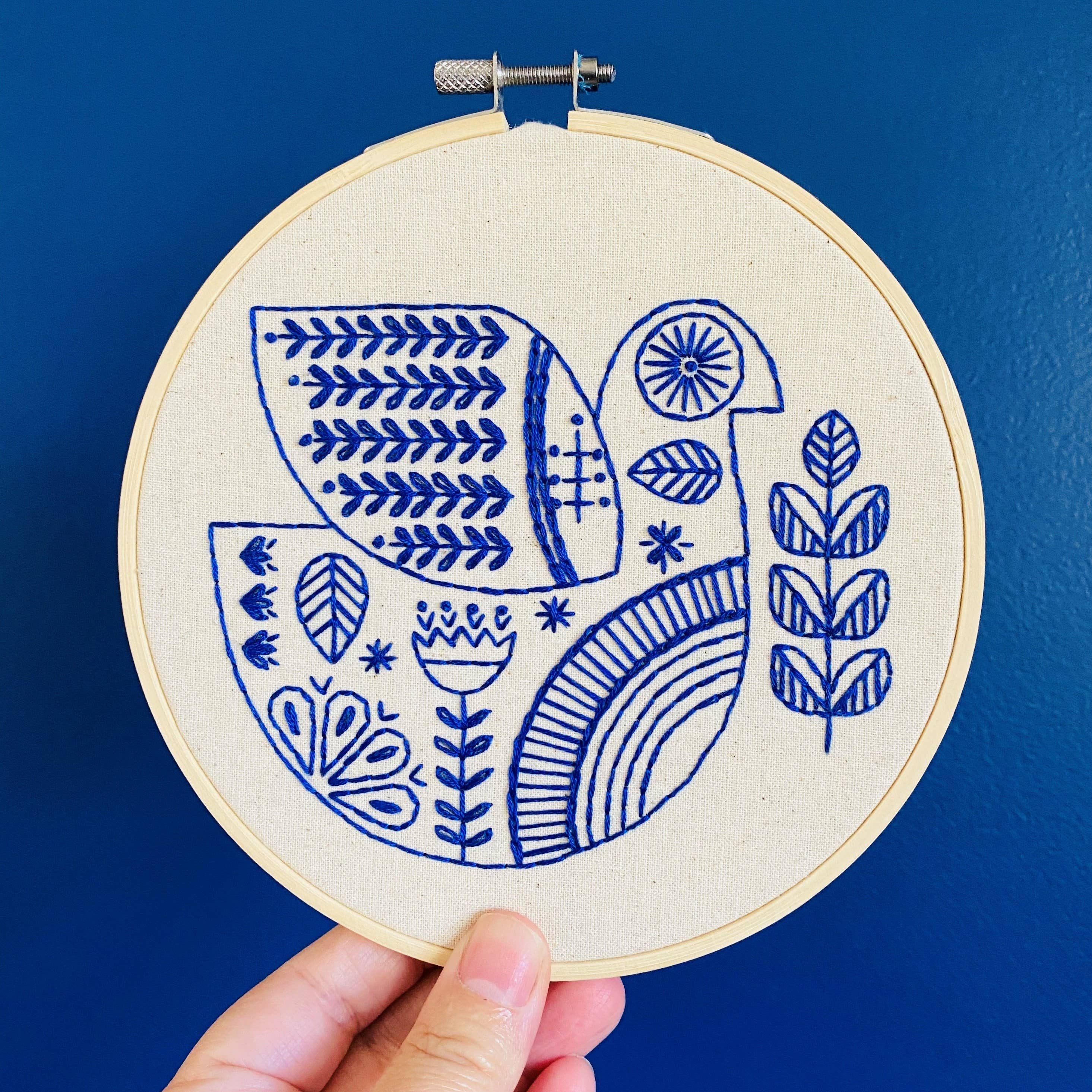 A hand holds the Hygge Dove Complete Embroidery Kit by Hook, Line & Tinker, featuring a blue and white stylized bird, leaves, and geometric patterns on an embroidery hoop against a solid blue background.