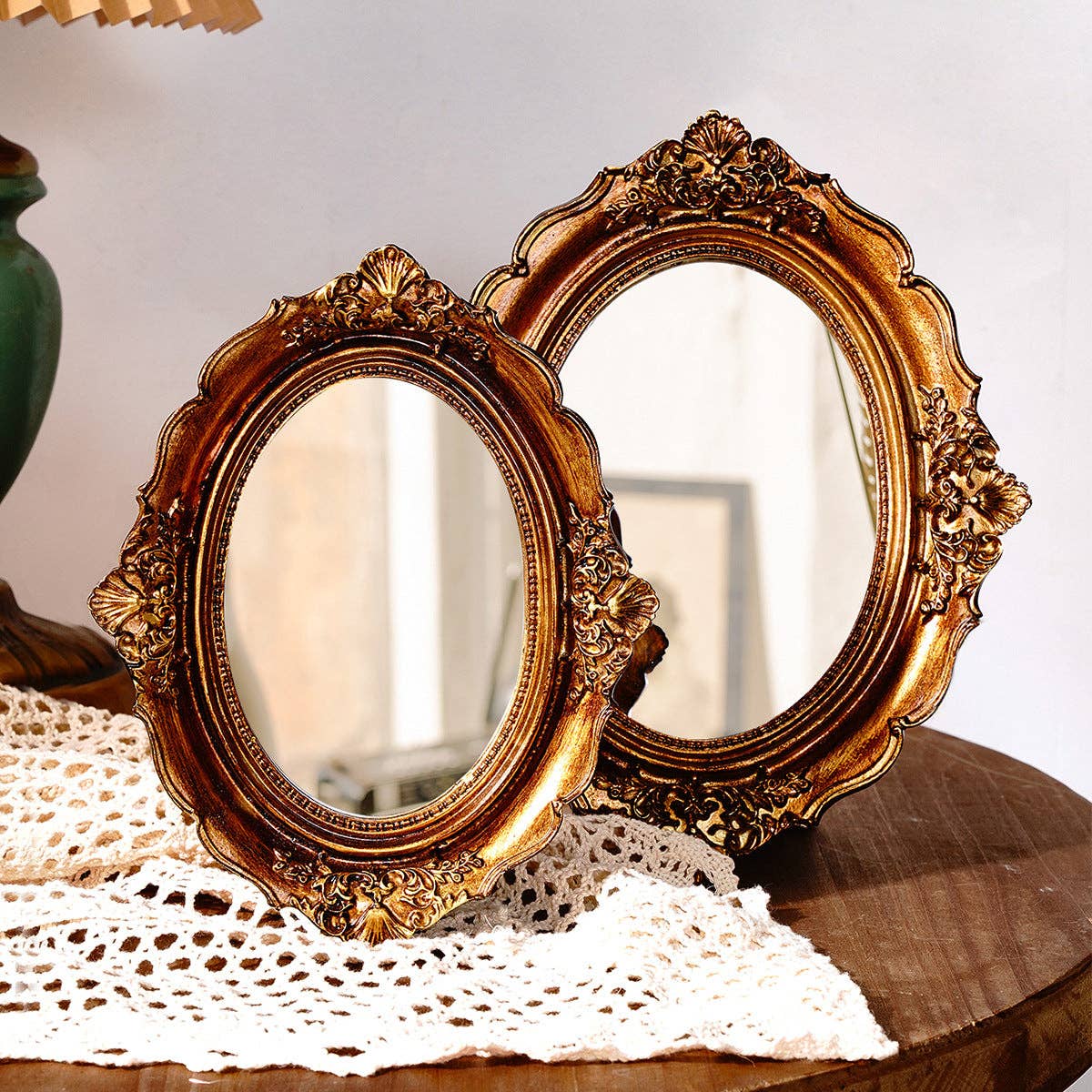 Two Vagabond Vintage "Vintage Style Ornate Oval Table Mirrors: Medium" with antique gold frames sit on a round table draped in white lace, creating an elegant, softly lit display that highlights their vintage charm.