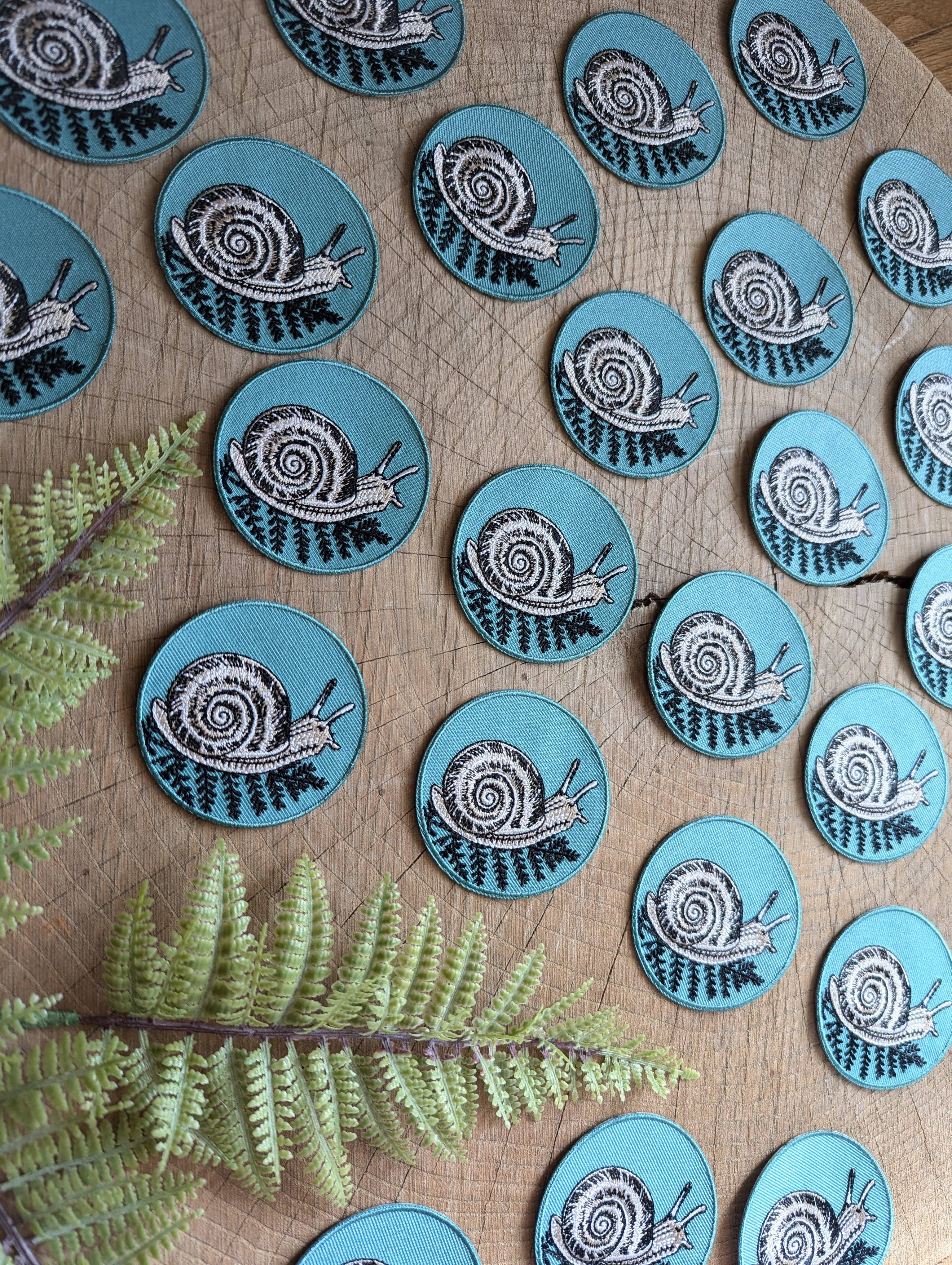 The ilikesara | art + goods 2.5" round teal iron-on patch features a snail with fern design, shown in rows on wood with green fern leaves for a cozy, nature-inspired flair—no backer card or packaging included.