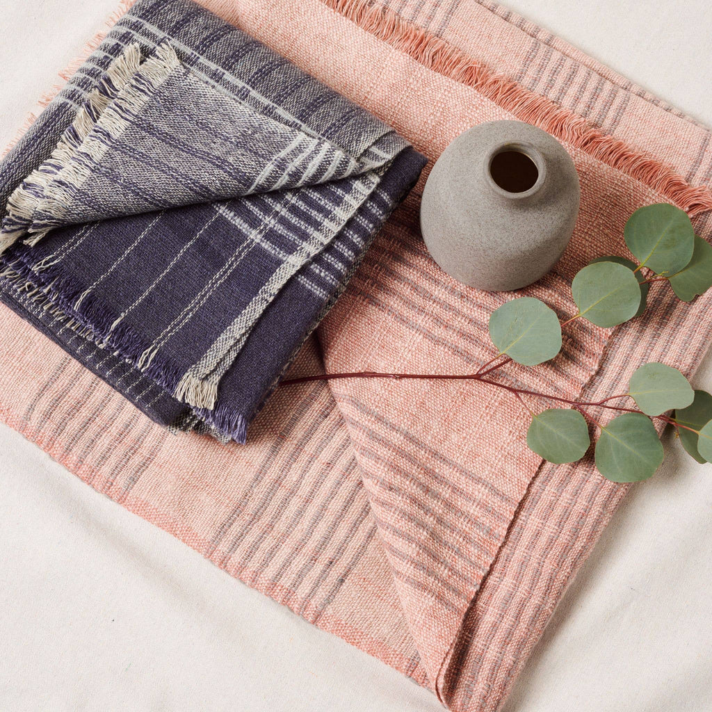 A folded Silica Rose Throw in shades of salmon gray stripes by Variously, along with a small gray vase and a branch with green leaves, are arranged on a light surface.