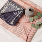 A folded Silica Rose Throw in shades of salmon gray stripes by Variously, along with a small gray vase and a branch with green leaves, are arranged on a light surface.
