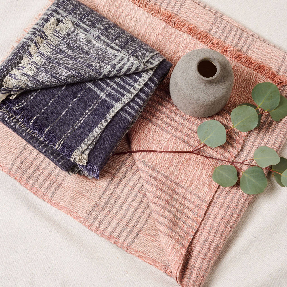 A folded Silica Rose Throw in shades of salmon gray stripes by Variously, along with a small gray vase and a branch with green leaves, are arranged on a light surface.